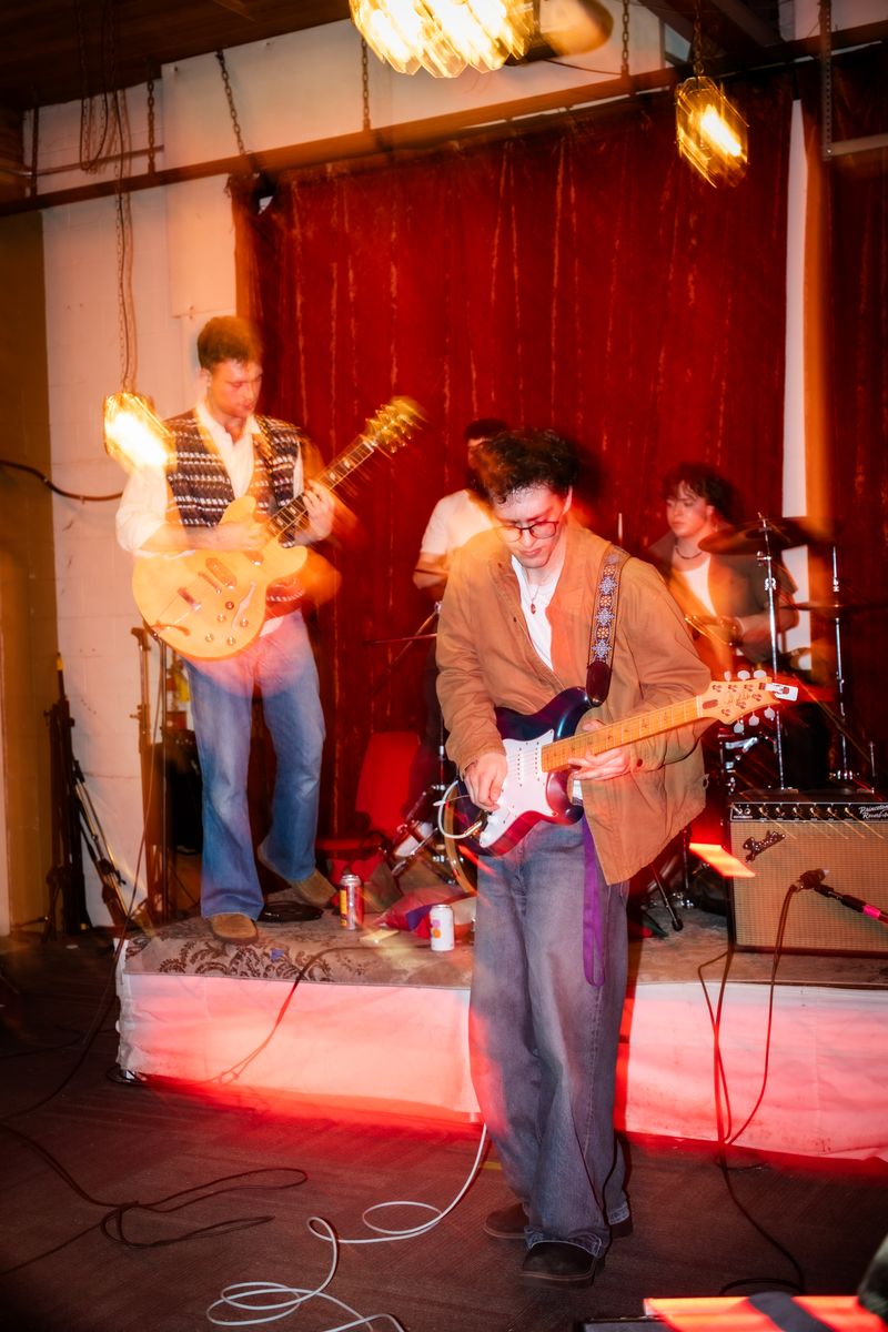 Guitarist in a corduroy jacket plays in front of a low stage while bandmates play behind him against a red velvet curtain