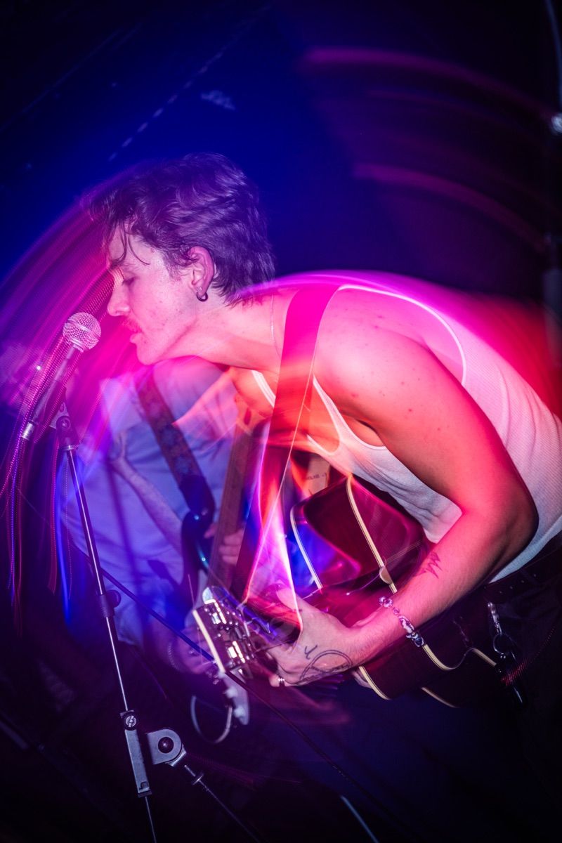 Singer in a white tank top leans into the microphone with eyes closed, playing guitar inside a swirl of purple and pink light