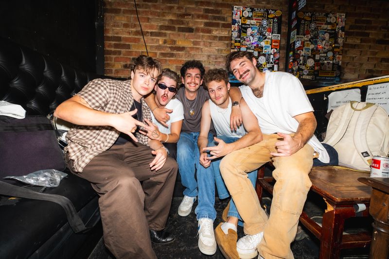 The five members of Dirty Aesthetic crowd onto a backstage couch by a brick wall of stickers, grinning and throwing hand signs