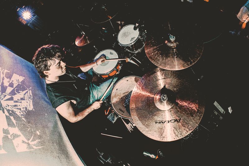 Drummer seen from directly above, sticks on the snare, with large copper cymbals filling the foreground of a dark stage