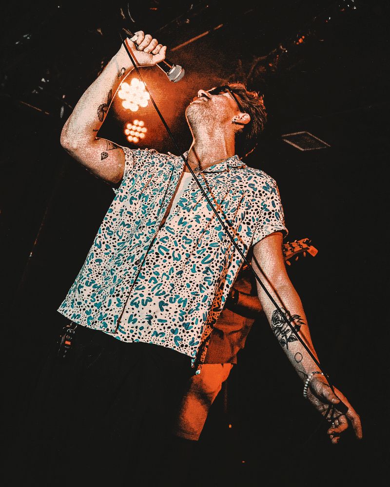 Shot from below, a singer in sunglasses and a patterned shirt holds the microphone high overhead under orange stage light