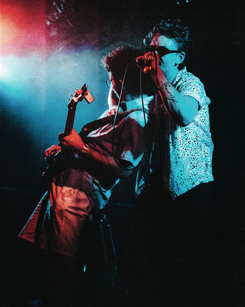 Singer in sunglasses and a spotted shirt grips the microphone beside a guitarist with curly hair, both lit in teal and red light