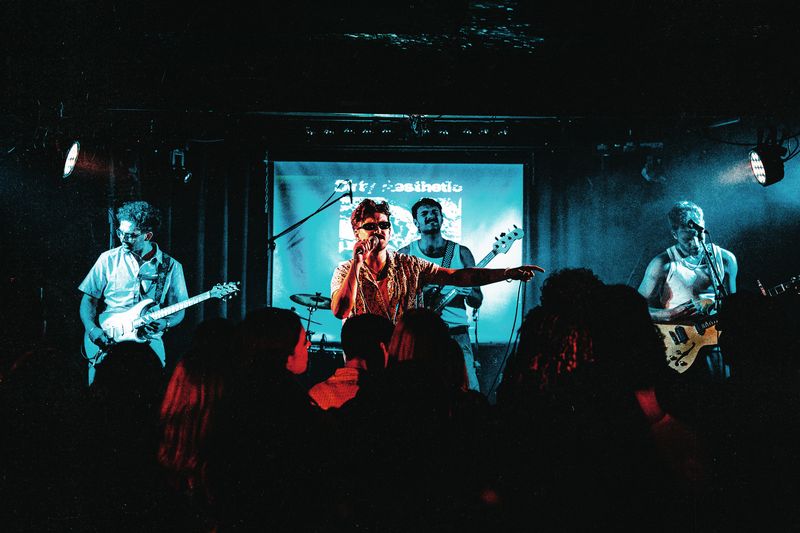 Dirty Aesthetic play under teal light with their name projected behind them as the singer points out over a silhouetted crowd