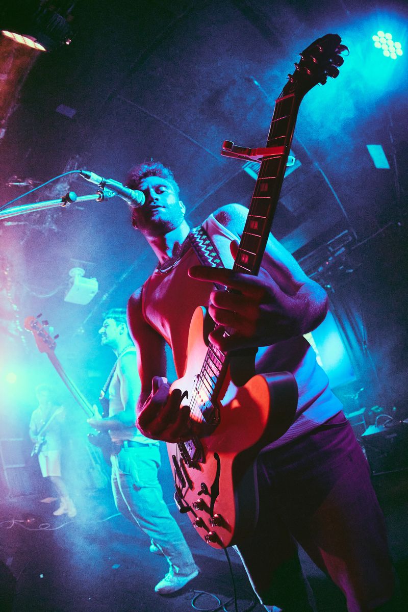 Low angle shot of a guitarist singing into a mic, red hollow body guitar thrust at the lens, two bandmates behind in blue haze.