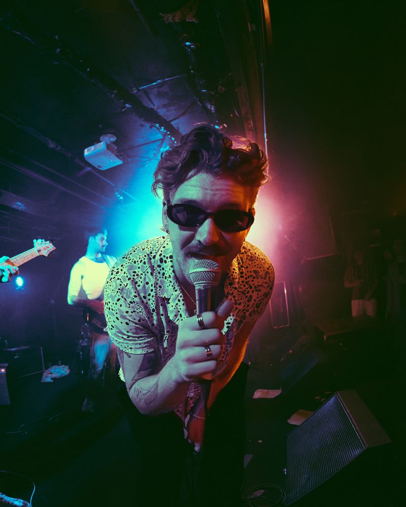 Singer in dark sunglasses and a spotted shirt leans into the lens gripping a mic, a guitarist glowing in blue haze behind.