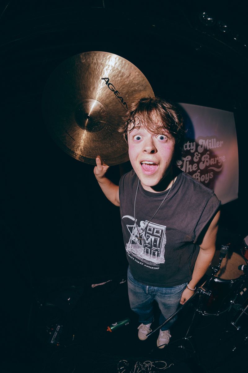 Wide eyed musician in a sleeveless tee gapes up at the camera, holding a cymbal beside his head next to the drum kit.