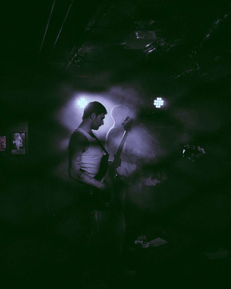Bassist in a white tank top plays in profile, lit from behind in violet haze with a ghostly silhouette at his shoulder.