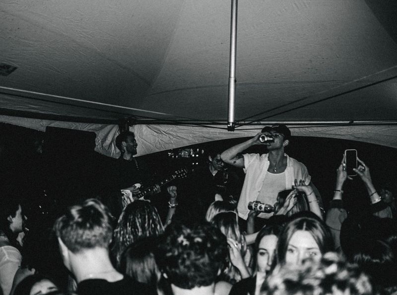 Singer in a white shirt swigs from a can under a tent canopy as a guitarist plays and fans hold up phones, black and white