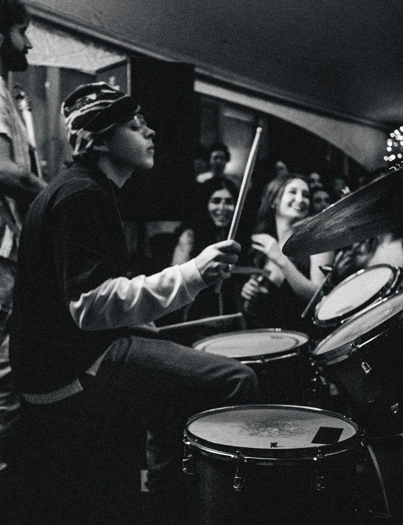 Drummer in a patterned bucket hat plays with eyes closed as smiling fans watch from behind the kit, black and white