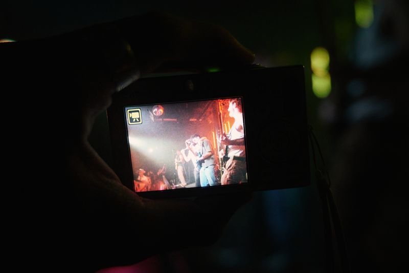 A hand holds a small camera whose screen shows the band playing under orange stage light, green bokeh in the dark behind.