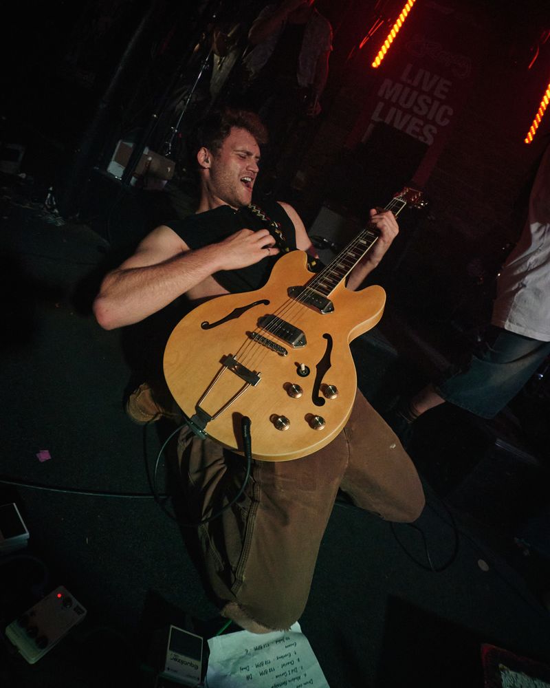 Guitarist in a black sleeveless shirt leans back mid shout with a blonde hollow body guitar, a Live Music Lives sign behind.