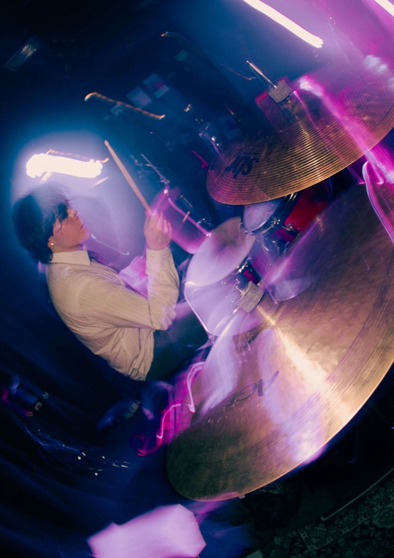Drummer in a cream striped shirt swings a stick behind large cymbals, blurred by purple and pink light trails