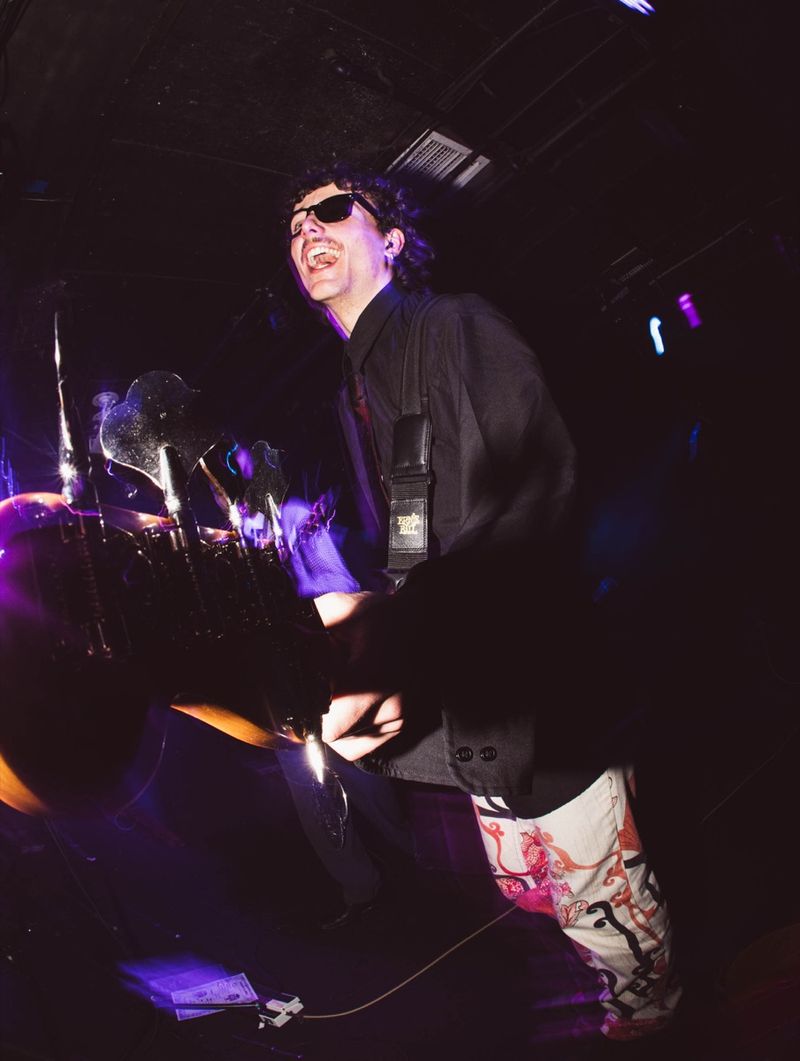 Guitarist in sunglasses and patterned trousers laughs mid song, lit purple under a low club ceiling