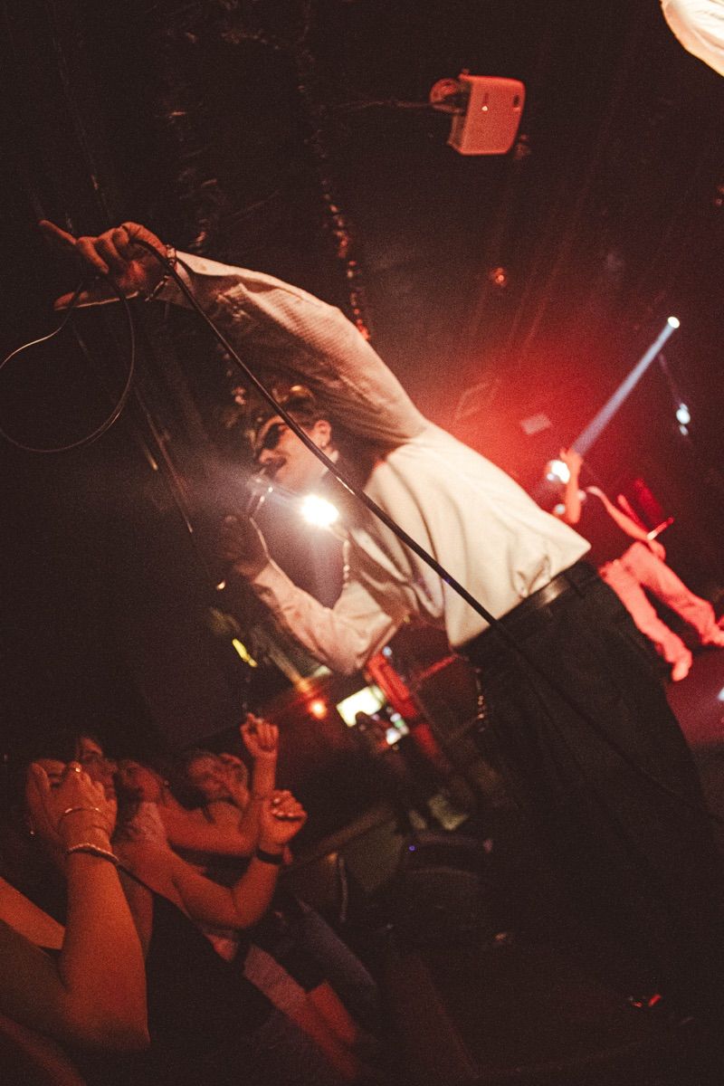 Singer in sunglasses and a white shirt leans over the front row, mic cable raised overhead, under warm red light