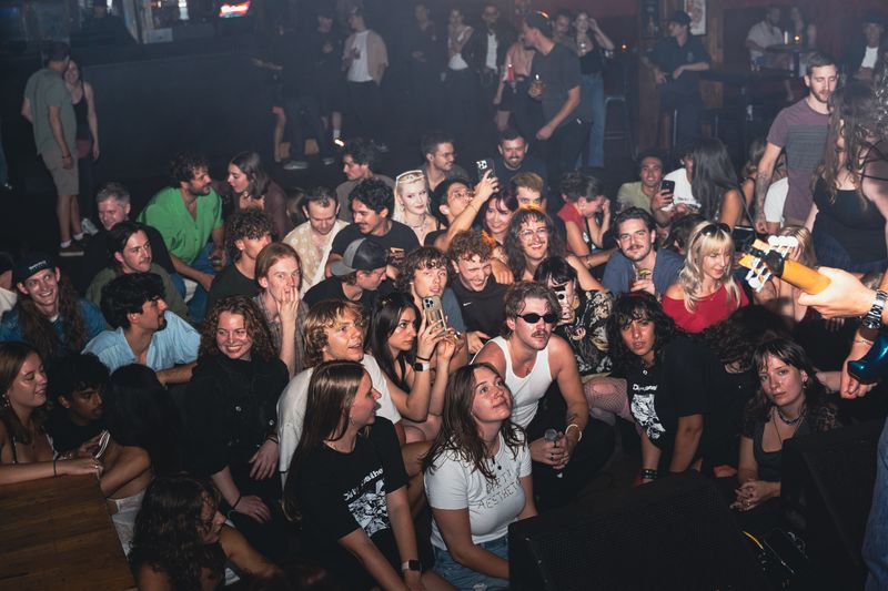 A packed crowd, front rows seated on the floor, smiles up at the stage, one fan wearing a Dirty Aesthetic shirt.
