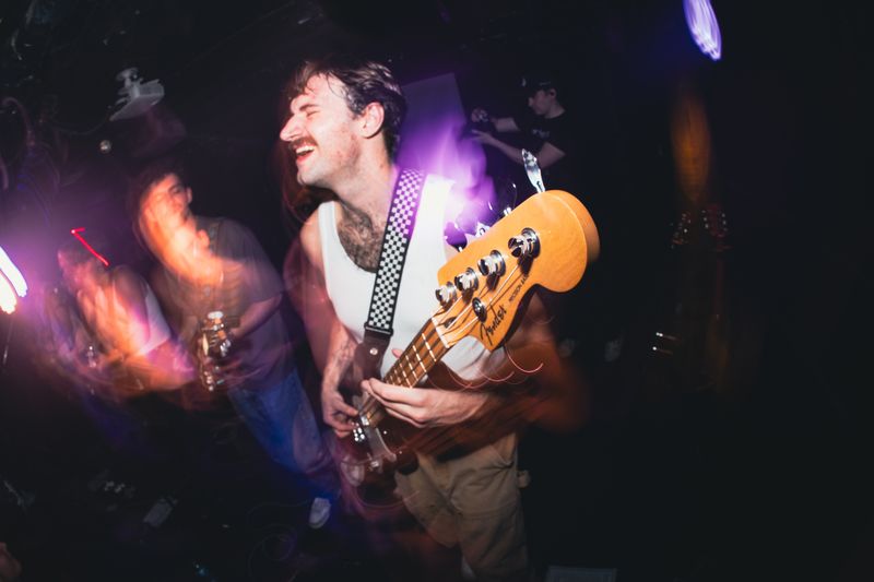 Mustached bassist in a white tank grins with eyes closed, Fender headstock thrust forward, amid purple and orange light trails.