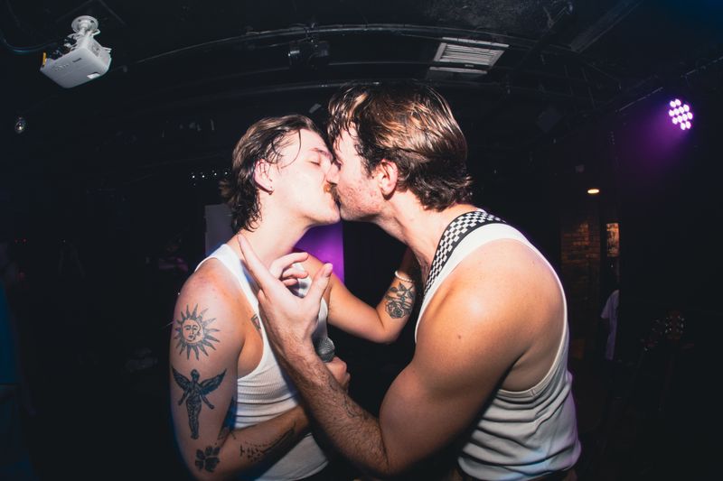 Two Dirty Aesthetic members in white tank tops kiss onstage, one holding a microphone, under a purple stage light