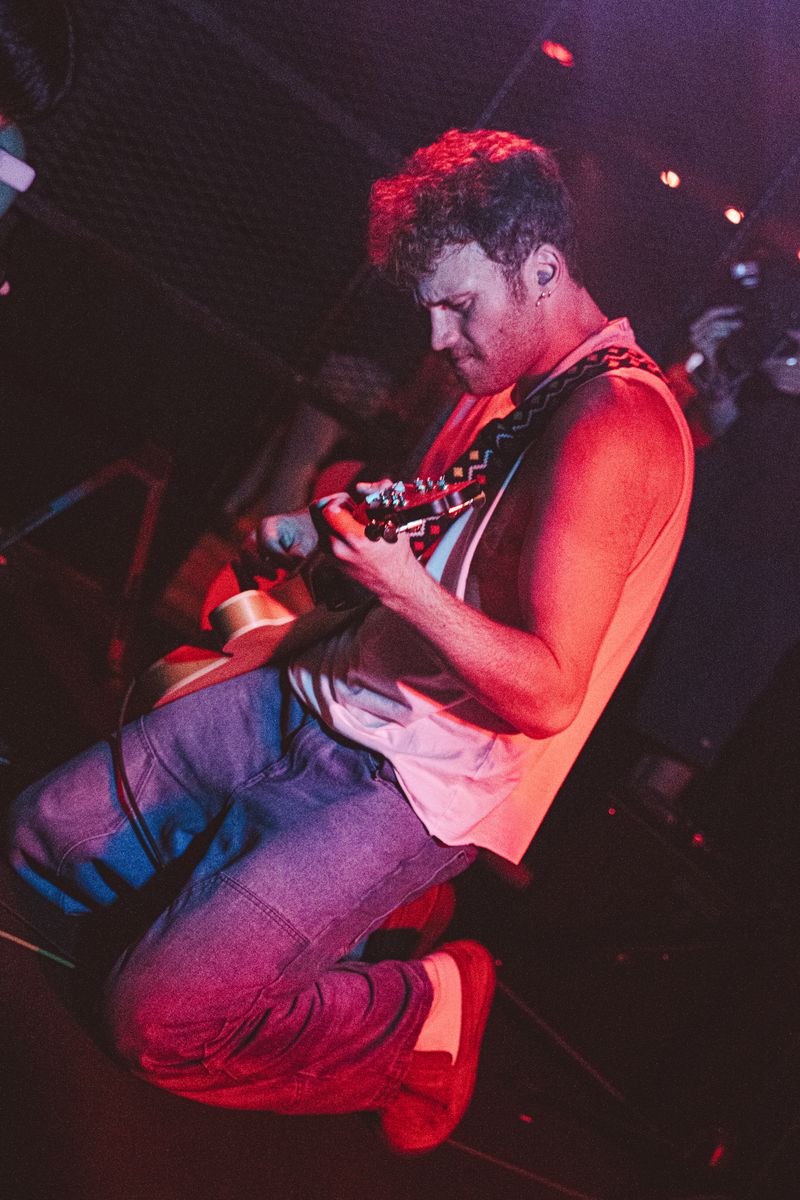Guitarist in a tank top and jeans crouches low on stage, head bowed over his guitar, washed in red light