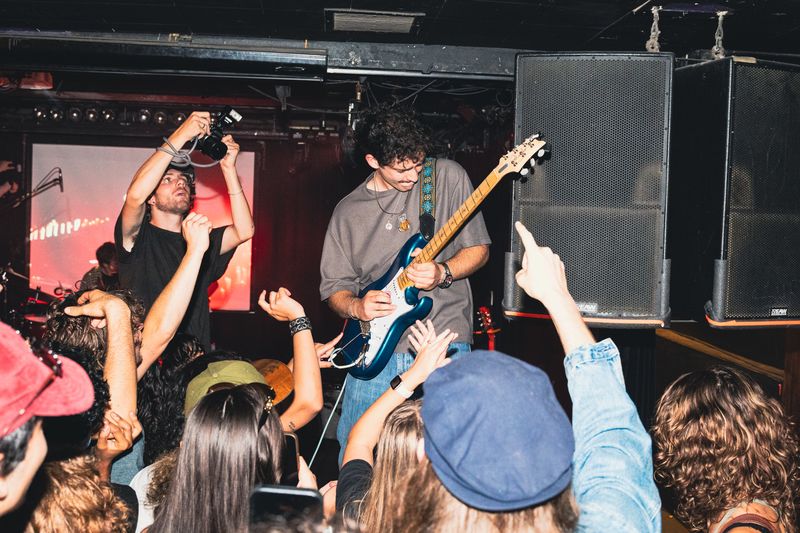 Guitarist with curly hair and a blue guitar looks down at raised hands as a photographer shoots from the stage beside him