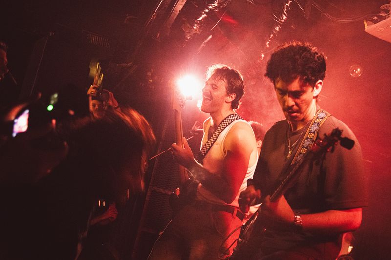 Two members play through red haze and a bright backlight, one shouting with eyes shut, as a fan films on a phone up front