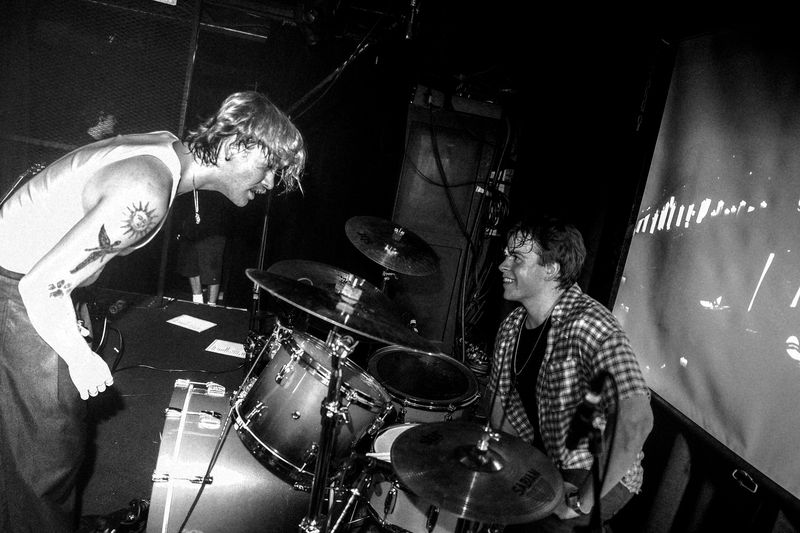 Tattooed band member in a white tank top leans over the drum kit toward the smiling drummer in a plaid shirt, black and white