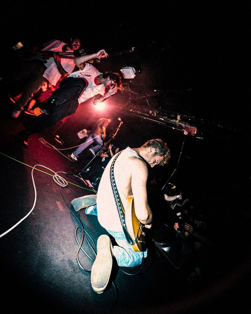 Guitarist in a white tank top kneels on stage under red light, bandmates leaning back behind him in a tilted wide shot
