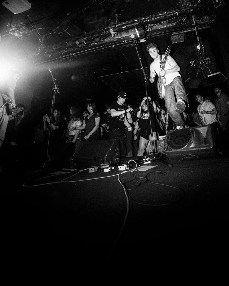 Guitarist in a white tank top kicks a leg up atop a stage monitor, the crowd dancing behind him, black and white