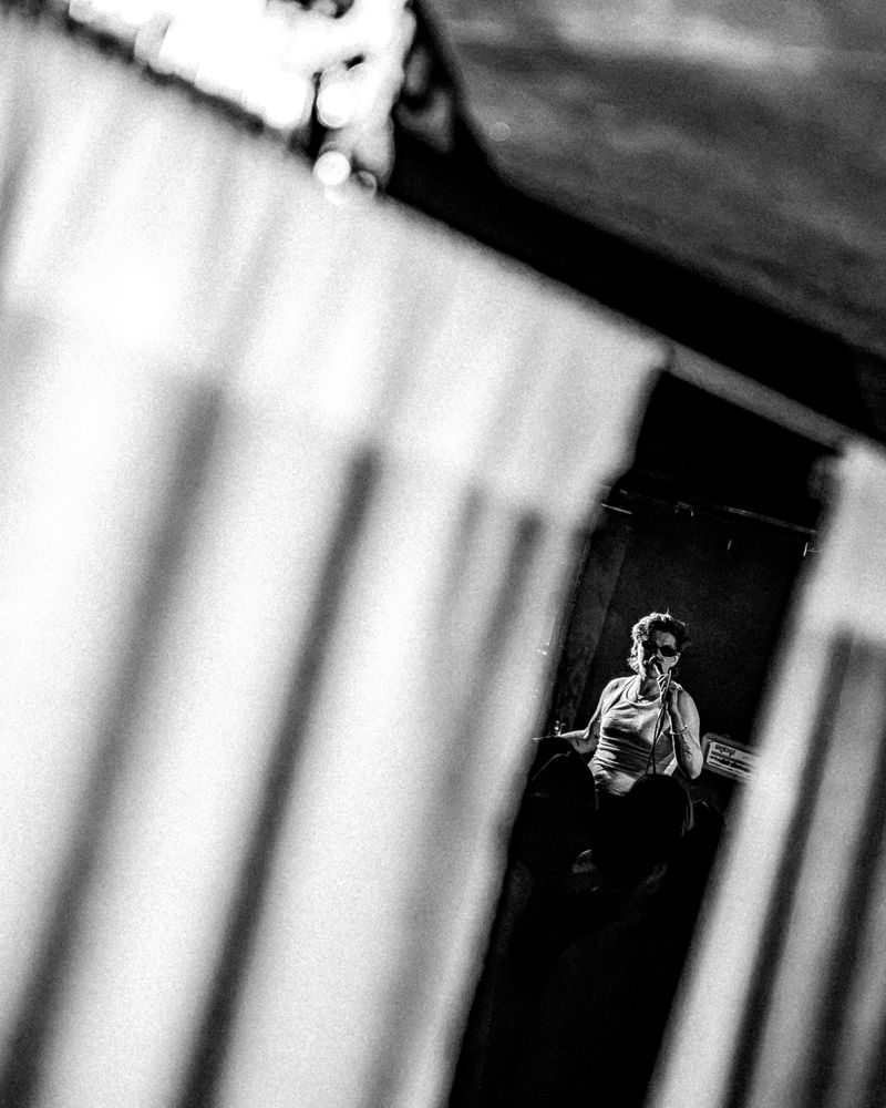 Singer in sunglasses sings into a microphone, glimpsed through a gap in blurred foreground shapes, black and white