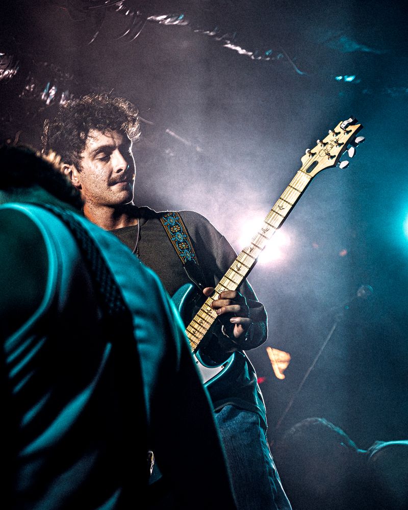 Guitarist with curly hair and a moustache plays under teal haze and backlight, a bandmate blurred in the foreground