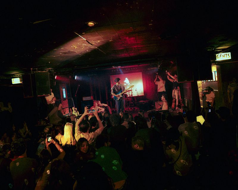 Fans hold up phones in a packed room under red light as Dirty Aesthetic plays in front of a glowing projection screen