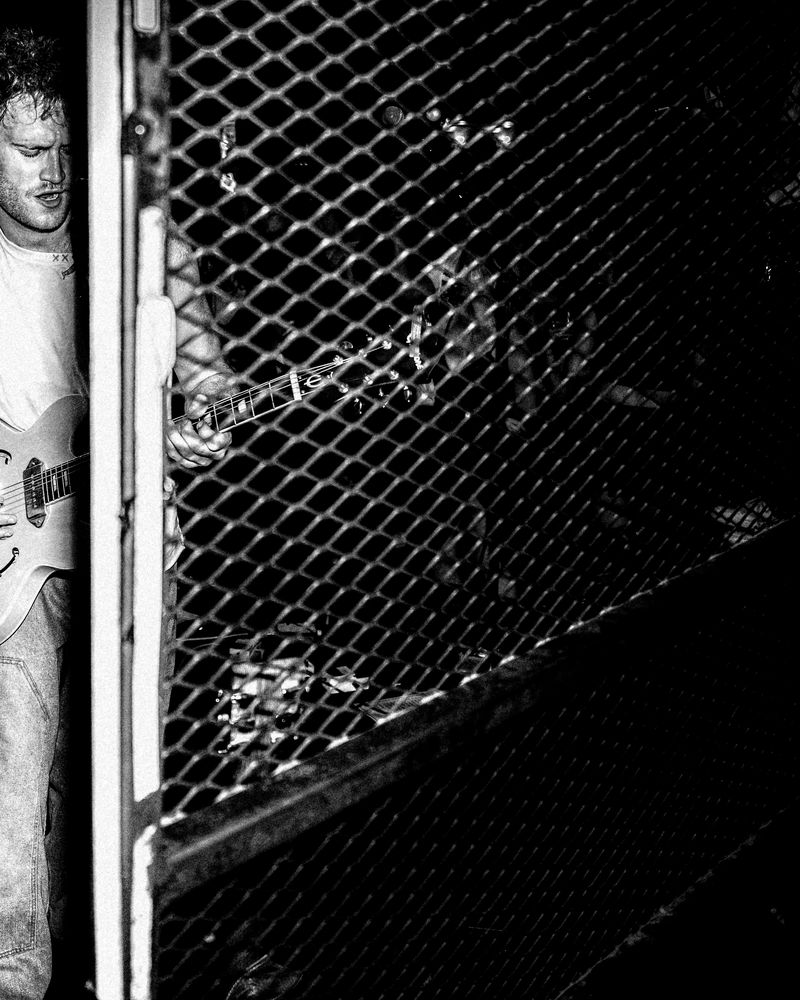 Guitarist in a white tee plays a hollow body guitar beside a wire mesh gate, with blurred figures behind the mesh, in black and white.
