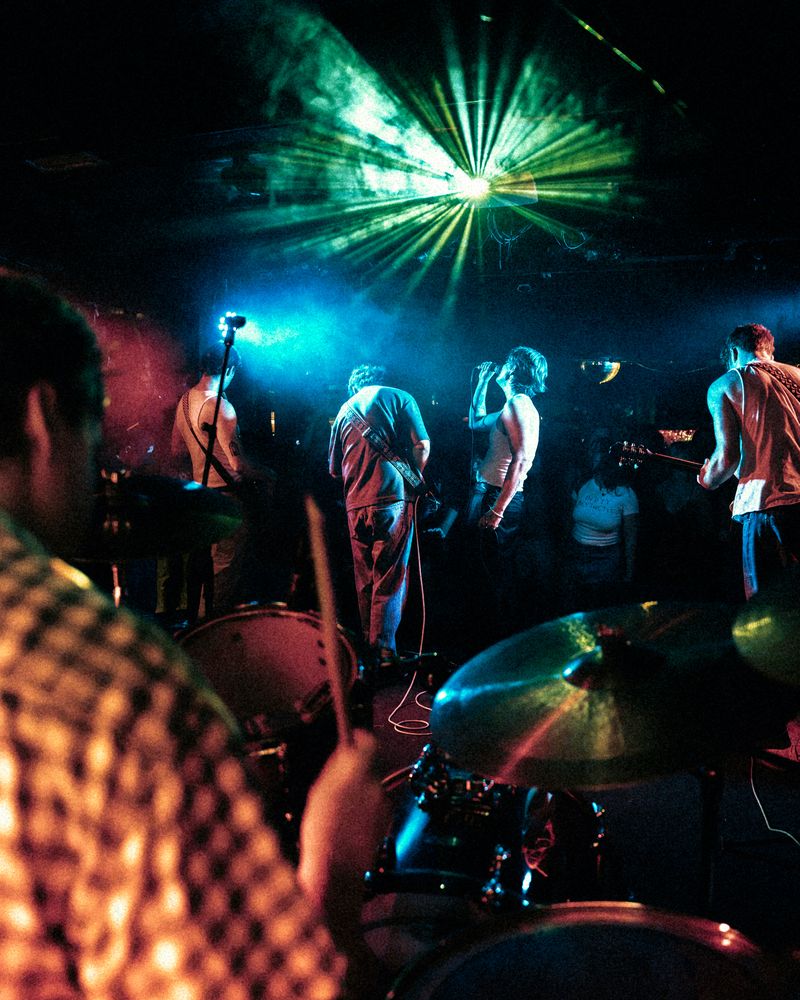 From behind the drum kit, the band faces the crowd under a green laser burst and teal haze, the drummer blurred in the foreground.