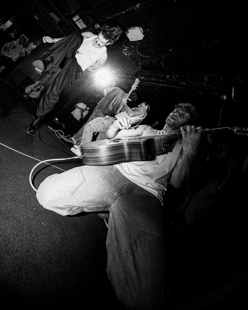 Guitarist leans back with a knee thrust at the lens, playing an acoustic guitar as two bandmates thrash behind him, in black and white.