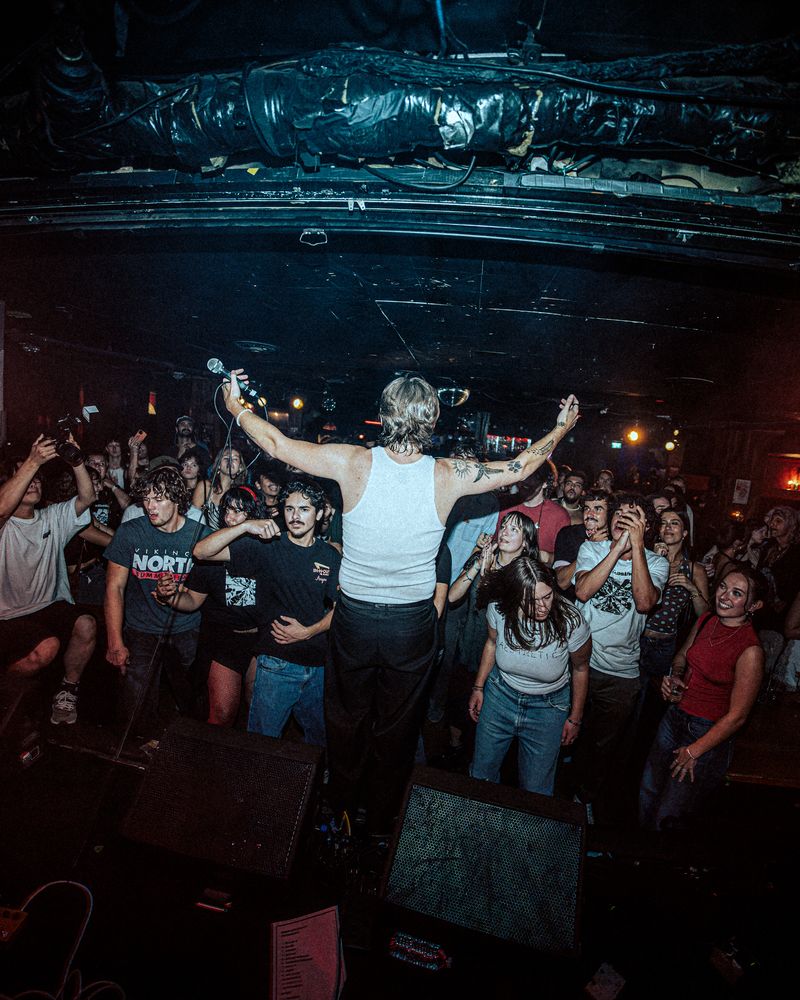 Singer in a white tank top holds a microphone aloft and spreads his arms to a packed, cheering crowd, seen from behind on stage.
